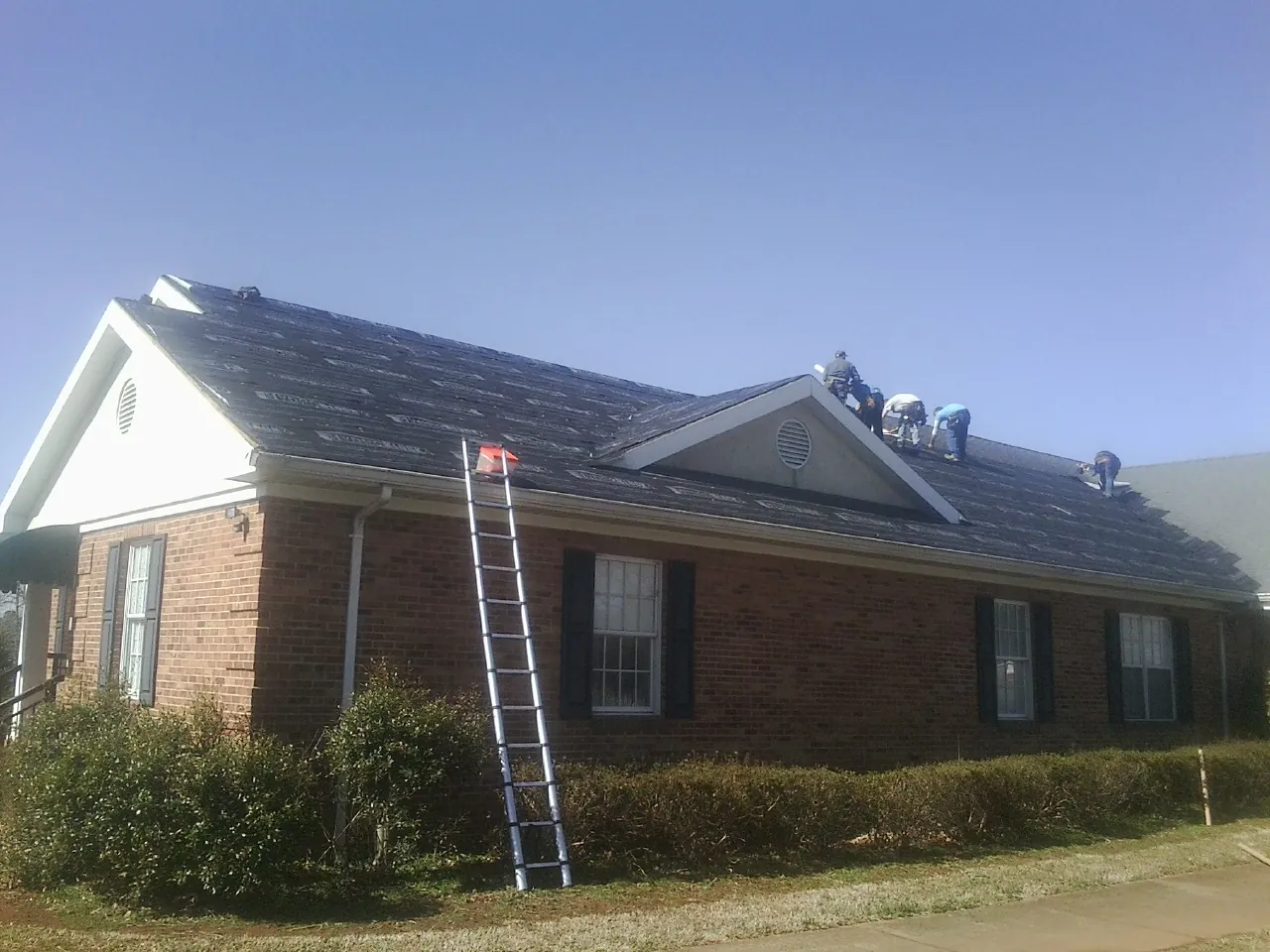 Crew tearing shingles off a brick ranch roof with a ladder set — Metal Roof Repair work by our crew in Eagle Bridge