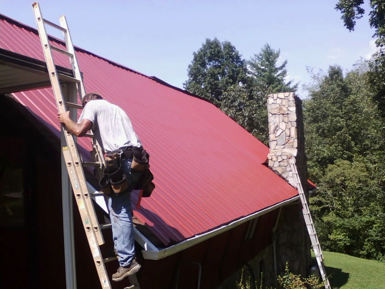 Roofer on a ladder beside a red metal roof and stone chimney — Soffit & Fascia Repair by Roofing Brothers in Eagle Bridge